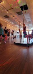 Group pole fitness class in an airy indoor studio — woman practicing on a chrome pole atop a round stage while classmates watch, wooden floors, hanging string lights and colorful laser dots on the ceiling.