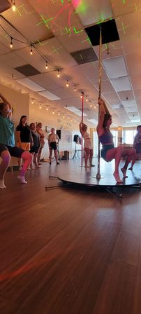 Group pole fitness class in an airy indoor studio — woman practicing on a chrome pole atop a round stage while classmates watch, wooden floors, hanging string lights and colorful laser dots on the ceiling.