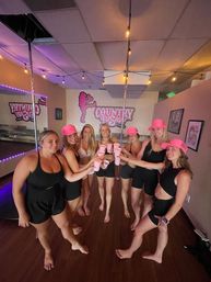 Seven women in black workout outfits and bright pink caps toasting with pink cups inside a pole-fitness studio with poles, string lights, mirrors and pink wall art.