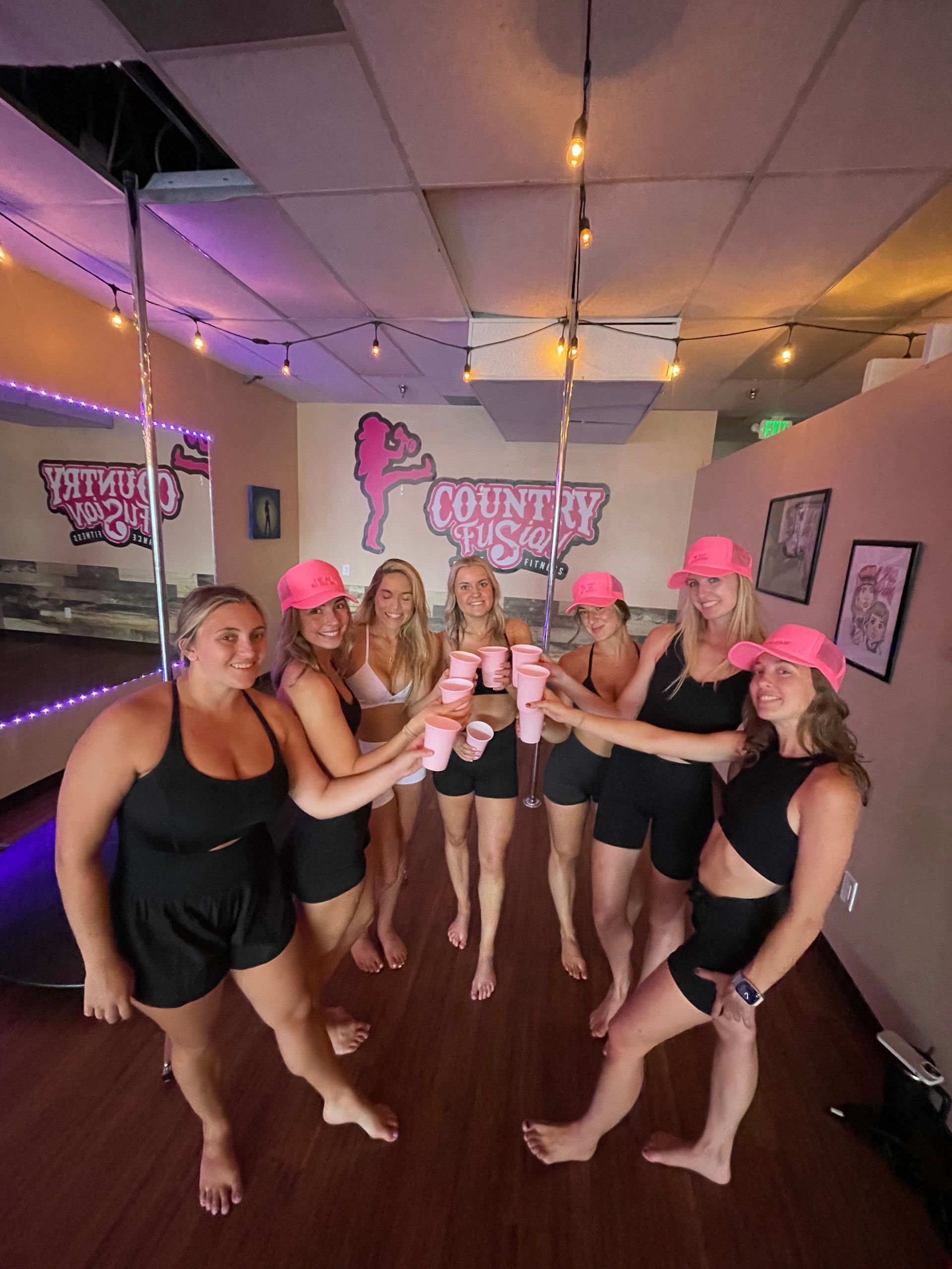 Group of women in athletic tops and shorts wearing matching pink caps, toasting with pink cups around a chrome pole in a pole-dance fitness studio lit by string lights and colorful accents.
