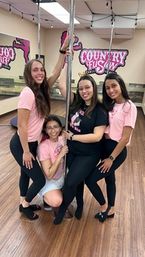 Four smiling women in matching pink shirts posing around a dance pole inside a bright pole-fitness studio with wood floors, mirrors and string lights — group fitness/dance class photo.