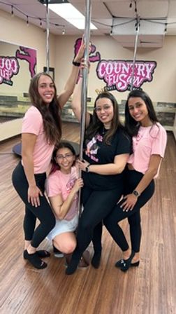 Five people posing around a chrome pole in a bright hardwood-floor pole dance studio with mirrors and string lights, wearing pink and black workout clothes.