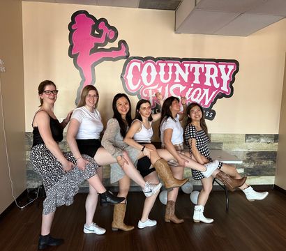 Six women lined up in a country-style dance studio kicking legs and smiling, wearing cowboy boots and casual outfits beneath a pink western logo on the wall.