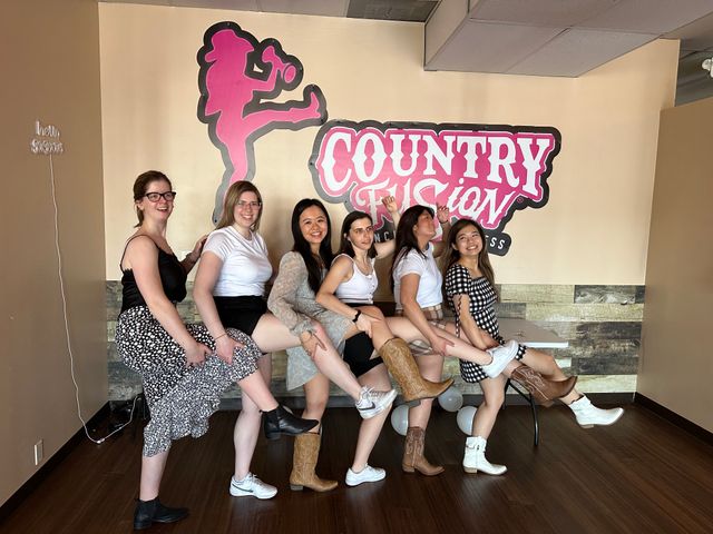 Six women lined up in a country-style dance studio kicking legs and smiling, wearing cowboy boots and casual outfits beneath a pink western logo on the wall.