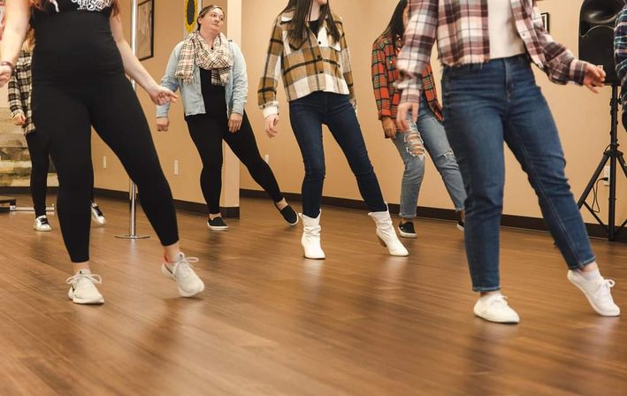Group of dancers mid-step, grooving in a casual indoor dance rehearsal at a wood‑floored studio, wearing jeans, leggings, flannels, boots and sneakers.