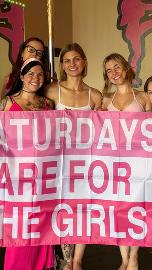 Eight women in pink outfits smiling in a dance studio around a pole, holding a large pink banner reading 'SATURDAYS ARE FOR THE GIRLS' — girls' day pole-fitness celebration.