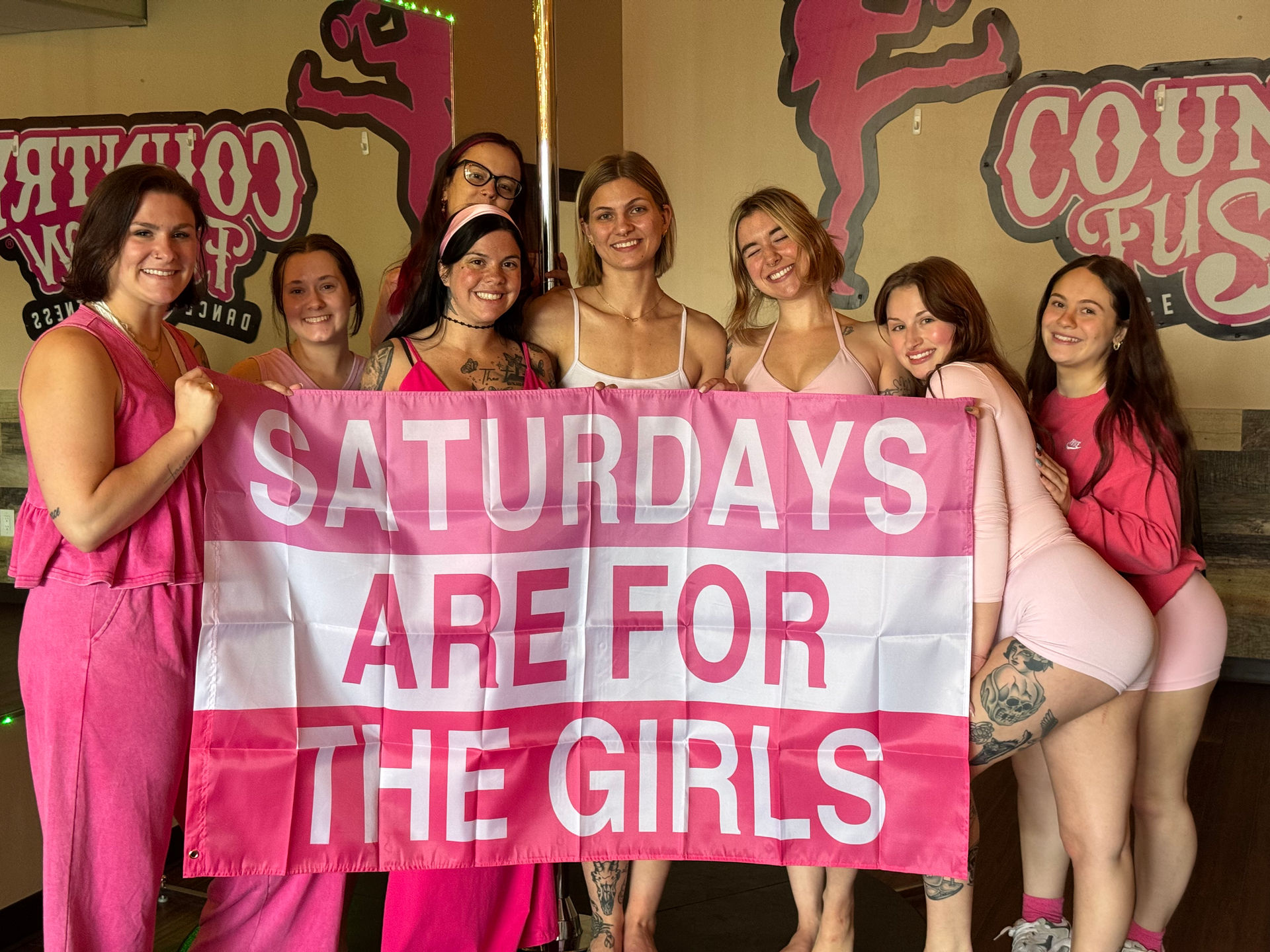 Eight women in pink outfits smiling in a dance studio around a pole, holding a large pink banner reading 'SATURDAYS ARE FOR THE GIRLS' — girls' day pole-fitness celebration.