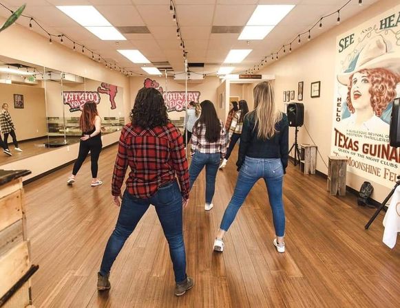 Group of women in jeans and plaid practicing country line dance in a bright studio with hardwood floors, mirrored wall, and vintage western poster.