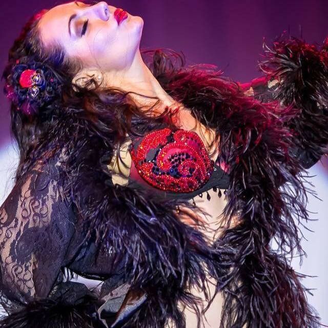 Burlesque performer in a red sequined bra and black lace sleeves draped in a black feather boa, striking a dramatic head-back pose on a purple-lit cabaret stage.