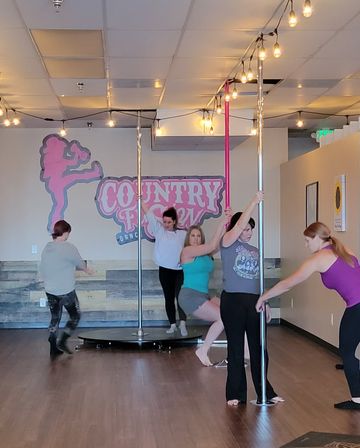 Women in a pole-fitness group class practicing spins and holds on chrome and pink poles in a bright dance studio with wood floors, string lights, and a playful mural backdrop.