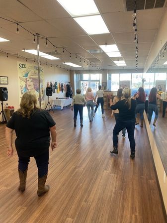 Women in cowboy boots taking a line-dance class in a sunlit storefront dance studio with wood floors, mirrored wall, string lights and speakers.