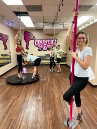 Women in a pole-fitness class in a wood‑floored indoor studio, smiling and posing on pink and silver poles in front of mirrors and a dancer silhouette mural.