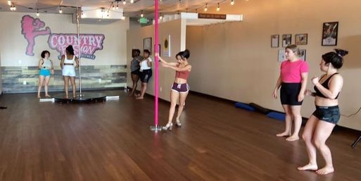 Group of women taking a pole fitness class in a bright dance studio with a pink pole, wall mirror, hardwood floor, string lights and casual workout attire.