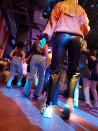 Low-angle shot of a vibrant nightclub dance floor with people dancing under colorful stage lights, focusing on legs in black leather pants, a pink sweater and white sneakers.