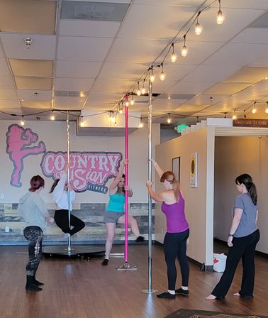 Women in a lively pole fitness class practicing spins and climbs on chrome and pink poles under string lights in an airy indoor studio