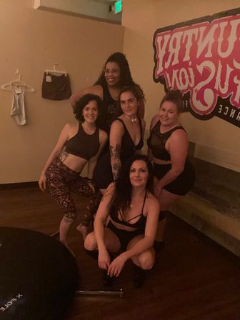 Five women in dancewear smiling and posing inside a dimly lit pole-dance studio with wooden floor, wall signage and a portable pole base