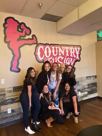 Seven women smiling and posing indoors at a country‑themed studio, in front of a large pink logo and kicking‑figure wall mural with rustic wood paneling.