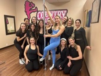 Group of smiling women in a pole fitness class inside a dance studio — one woman in blue performing a pole pose while classmates in black gather on a hardwood floor under a pink wall mural.