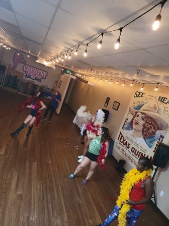Women rehearsing a burlesque-style dance in an indoor wood‑floored dance studio, wearing colorful feather boas and casual workout clothes beneath hanging string lights and a vintage cowgirl mural.