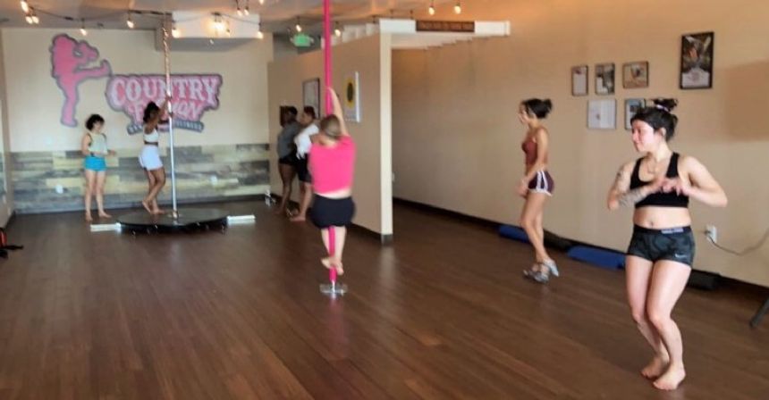 Group pole fitness class in a bright indoor dance studio — women in athletic wear practicing pole and floor dance on a wood floor with studio wall art.