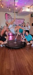 Seven women in a bright indoor pole-fitness studio posing around a dance pole — one woman in white holding a mid-air pose while friends kneel and react playfully under string lights and colorful ceiling effects, wood floor and studio logo on the wall.