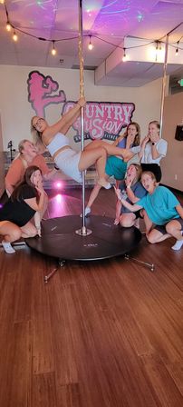 Seven women in a bright indoor pole-fitness studio posing around a dance pole — one woman in white holding a mid-air pose while friends kneel and react playfully under string lights and colorful ceiling effects, wood floor and studio logo on the wall.