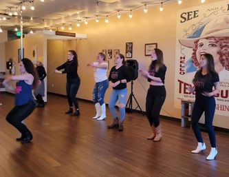 Women in a line-dance class practicing on a wood-floor studio under string lights, wearing jeans and cowboy boots in front of a vintage western mural.