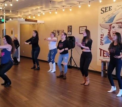 Women in a line-dance class practicing on a wood-floor studio under string lights, wearing jeans and cowboy boots in front of a vintage western mural.