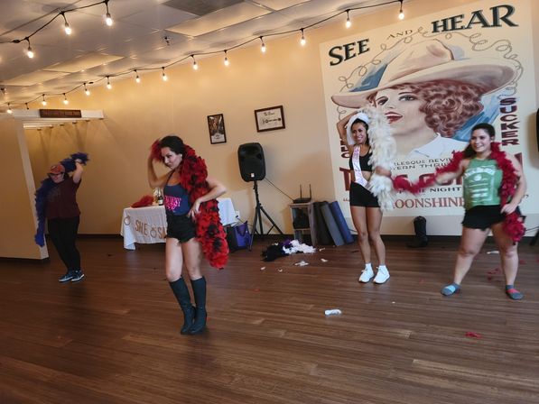 Four women rehearsing a playful dance in a studio with hardwood floors and string lights, wearing colorful feather boas and a sash, with a vintage cowgirl mural and speaker along the wall.
