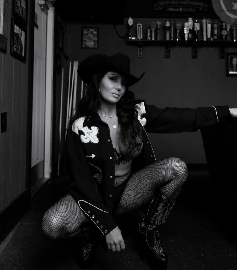 Black-and-white portrait of a cowgirl in a cowboy hat, embroidered western shirt, fishnet tights and cowboy boots squatting in a dim country bar with beer taps on the wall.