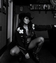 Black-and-white portrait of a cowgirl in a cowboy hat, embroidered western shirt, fishnet tights and cowboy boots squatting in a dim country bar with beer taps on the wall.