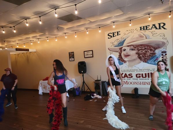 Bachelorette dance class practicing with colorful feather boas in a wood-floor studio under string lights, large vintage western poster mural on the wall.