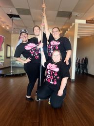 Four smiling people in matching black-and-pink 'LET'S GO GIRLS' shirts pose around a shiny chrome pole in a bright pole-fitness studio with wood floors.