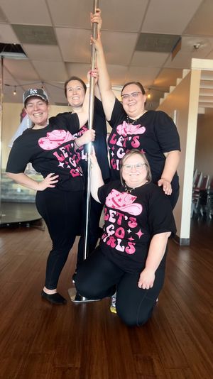 Four smiling people in matching black-and-pink 'LET'S GO GIRLS' shirts pose around a shiny chrome pole in a bright pole-fitness studio with wood floors.