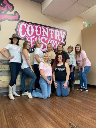 Eight women smiling and posing for a group photo inside a country-style dance studio beneath a large pink logo mural, wearing jeans, boots, and casual tees.
