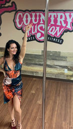 Smiling woman in a colorful orange-and-blue dress and strappy heels poses with arm raised beside a chrome pole in an indoor pole-fitness studio with wood floors and a pink wall mural.