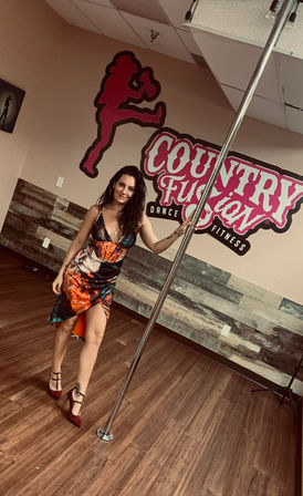 Woman in colorful dress and heels posing by a pole in a dance fitness studio with a pink mural and wood-plank floor