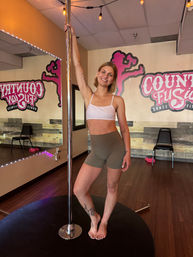 Smiling woman in a sports bra and shorts posing barefoot on a pole in a bright indoor pole-fitness dance studio with mirrors, wall graphics and wood floors.