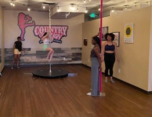 Group pole dance class in a bright studio with wood floors and string lights; a woman climbs a chrome pole on a circular platform while three others watch near a pink pole beneath a playful silhouette mural.