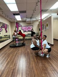 Five women smiling and posing on pink poles in a bright pole-fitness studio with wood floors and mirrored wall.