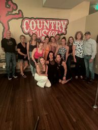 Cheerful mixed-age group posing for a photo in a country-themed dance studio with hardwood floor and a large red wall sign