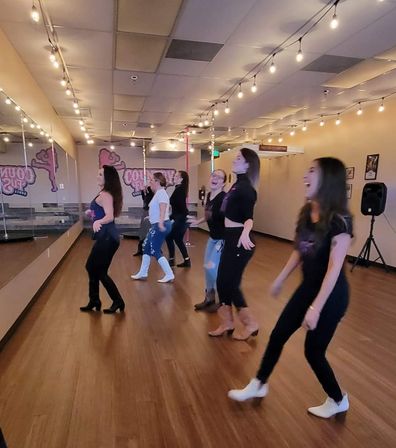 Women enjoying a line-dance class in a mirrored indoor dance studio with wood floors and string lights, laughing and dancing in boots and heels.