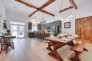 Open-concept modern farmhouse kitchen and dining room with exposed wooden ceiling beams, rustic wooden dining table and bench, island with barstools, hanging pendant lights, and sliding glass doors to a snowy backyard