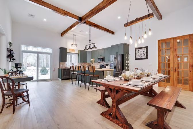Bright open-concept farmhouse kitchen and dining room with exposed wooden beams, long rustic wood dining table and benches set for a meal, green cabinetry island with bar stools, hanging Edison bulb pendant lights, stainless-steel appliances and sliding glass doors opening to a snowy backyard.