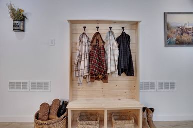 Rustic farmhouse entryway mudroom with light wood bench and shiplap coat rack holding four plaid jackets, wicker baskets and cowboy boots