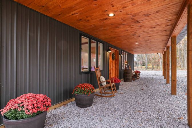 Rustic covered country porch with wood ceiling and posts, dark metal siding, gravel floor, rocking chair, barrels and potted red mums against a wooded autumn backdrop