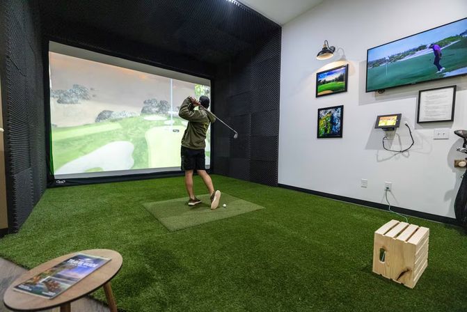 Indoor golf simulator room with artificial turf and hitting mat; a golfer swings toward a large projected virtual fairway, with a wall-mounted TV, framed golf prints and a small wooden stool nearby.
