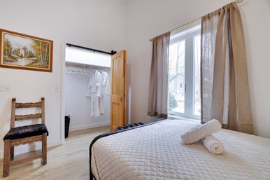 Inviting sunlit cottage-style bedroom with quilted bed and rolled towels, large window with beige curtains overlooking a yard, open closet with hanging white robes, wooden chair, framed landscape painting and light hardwood floors.