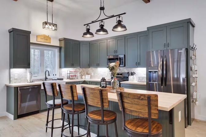 Cozy modern farmhouse kitchen with gray shaker cabinets, light wood island and four rustic wooden-and-metal barstools, stainless steel appliances, black industrial pendant lights, and tiled backsplash.