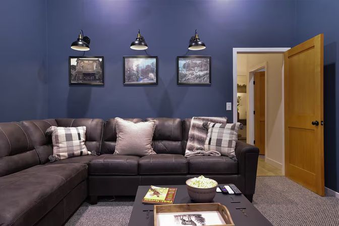 Cozy living room with navy blue wall, dark leather sectional sofa with plaid and soft pillows, three framed prints under industrial wall lamps, open wooden door, and coffee table holding a popcorn bowl and remotes — movie-night ready.